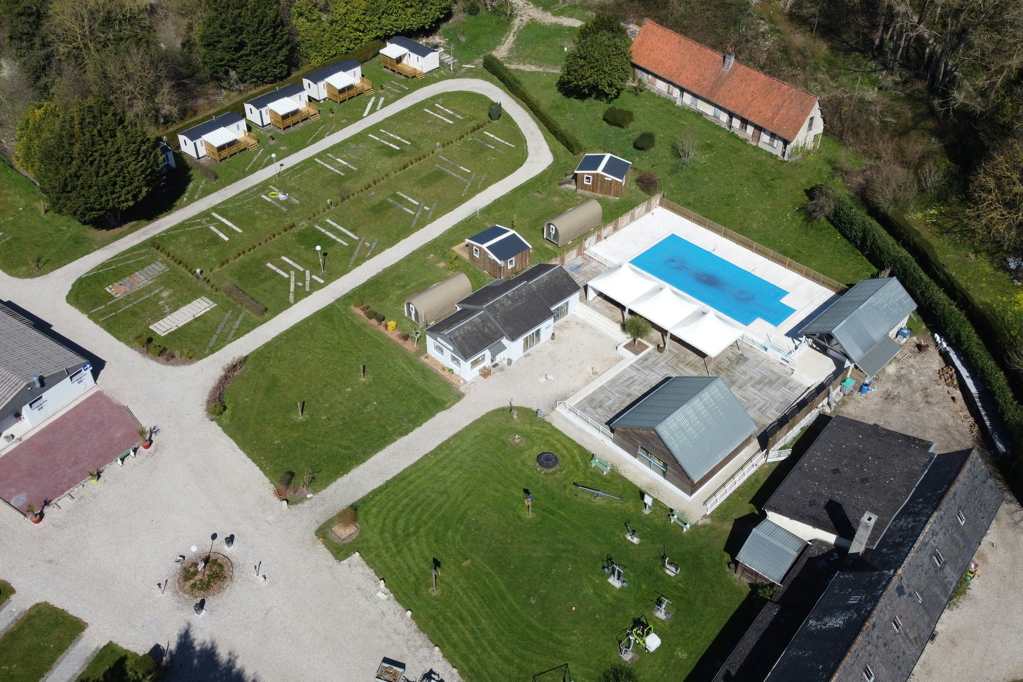 Luftaufnahme eines Campingplatzes mit Stellplätzen, Wohngebäuden und großem Pool im Garten, umgeben von Wiesen und Wegen.