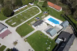 Luftaufnahme eines Campingplatzes mit Stellplätzen, Wohngebäuden und großem Pool im Garten, umgeben von Wiesen und Wegen.