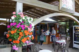 Blumen vor einer überdachten Terrasse am Campingplatz, Holzdach und Sitzgelegenheiten, im Hintergrund ein kleiner Kiosk.