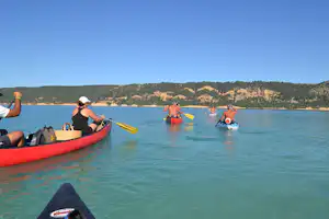 Rote Schlauchboote mit Paddlern im ruhigen See, im Hintergrund bewaldete Hügel und ein klarer Himmel.