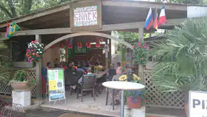 Snack-Bereich am Campingplatz mit überdachtem Eingang, Tischen und Stühlen, Blumenampeln und Fahnen im Hintergrund.