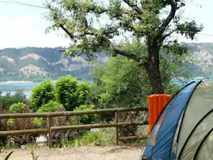 Campingzelt am Zaun mit Blick auf den See, im Vordergrund ein Baum und Büsche, dahinter bewaldete Hügel.
