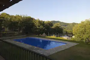 Schwimmbecken auf dem Campingplatz mit Blick auf grüne Hügel, Bäume und einen Weg am Rand.