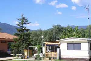 Ein weißes Mobilheim auf einem Campingplatz mit Holzzaun, Bäumen und Bergen im Hintergrund.