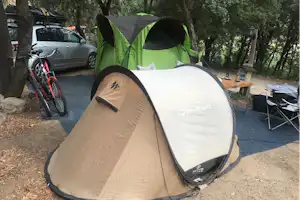 Grünes und beiges Campingzelt auf einem Stellplatz, daneben Fahrrad, Auto und Tisch mit Stühlen im Schatten der Bäume.