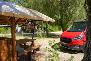 Rote SUV steht neben einem Holzunterstand mit Tisch und Bänken auf einem Kiesweg, umgeben von Bäumen und Grün.