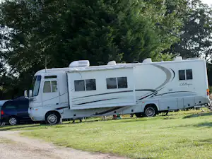 Wohnmobil auf einem Campingplatz am Grasrand, daneben ein Auto, im Hintergrund Bäume und Hecke.