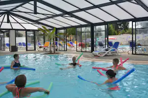 Kinder und Erwachsene im Schwimmbecken mit Schwimmhilfen unter einem Glasdach, Liegen und Sonnenschirme am Beckenrand.