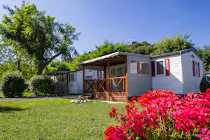 Mobilheim mit überdachter Terrasse, Holzgeländer und großen Fenstern, im Vordergrund rote Blumen und grüner Rasen.