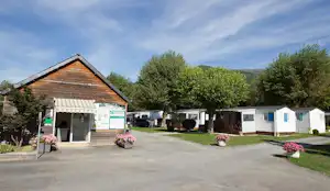 Eingang mit Holzgebäude, Markise und Infotafel, daneben Wohnwagen und Stellplätze, gepflasterter Weg, Blumenbeete und Bäume.