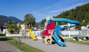 Spielplatz mit Rutsche und Kletterelementen auf dem Campingplatz, im Hintergrund Wohnwagen und Gebäude, Berge und Bäume.