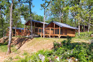 Holzhütten mit überdachter Terrasse im Wald, Holzgeländer und Zäune vor den Gebäuden, umgeben von Bäumen.