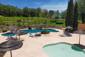 Poolbereich mit Sonnenschirmen, Liegestühlen und Becken auf einer Terrasse, dahinter Rasen, Bäume und Spielplatz im Campinggelände.