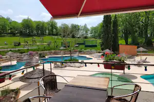 Campingpool mit türkisfarbenem Wasser, Liegen und Sonnenschirmen auf der Terrasse, im Hintergrund Spielgeräte und viel Grün.