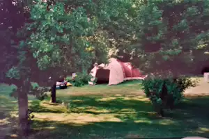 Zelt auf einer grünen Wiese im Campingbereich, umgeben von Bäumen und Sträuchern, mit Sitzbank und Tisch in der Nähe.