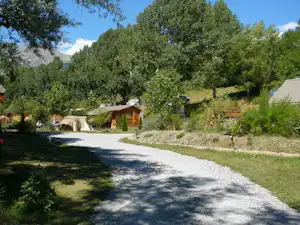 Schotterweg auf einem Campingplatz mit Zelt, kleinen Bäumen und einem Holzhaus im Hintergrund.