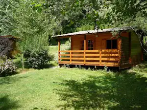Holzhütte mit Veranda und Holzgeländer auf einer grünen Wiese, umgeben von Bäumen und Sträuchern.