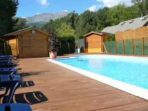 Schwimmbecken auf Holzdeck mit Liegestühlen, daneben Holzsauna und Sichtschutzzaun, umgeben von Bäumen und Bergen.