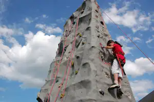 Kletterwand mit Klettergurt und Seilen, ein Kind klettert nach oben vor blauem Himmel mit Wolken.