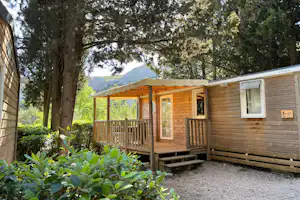 Holzbungalow mit Terrasse und Stufen am Campingplatz, umgeben von Bäumen und Sträuchern, mit Blick auf die Berge.
