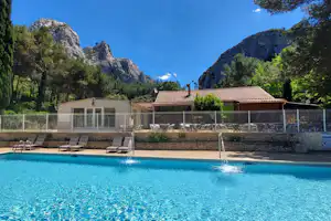 Schwimmbad eines Campingplatzes mit Liegen am Beckenrand, dahinter Gebäude und Berge in der Ferne.