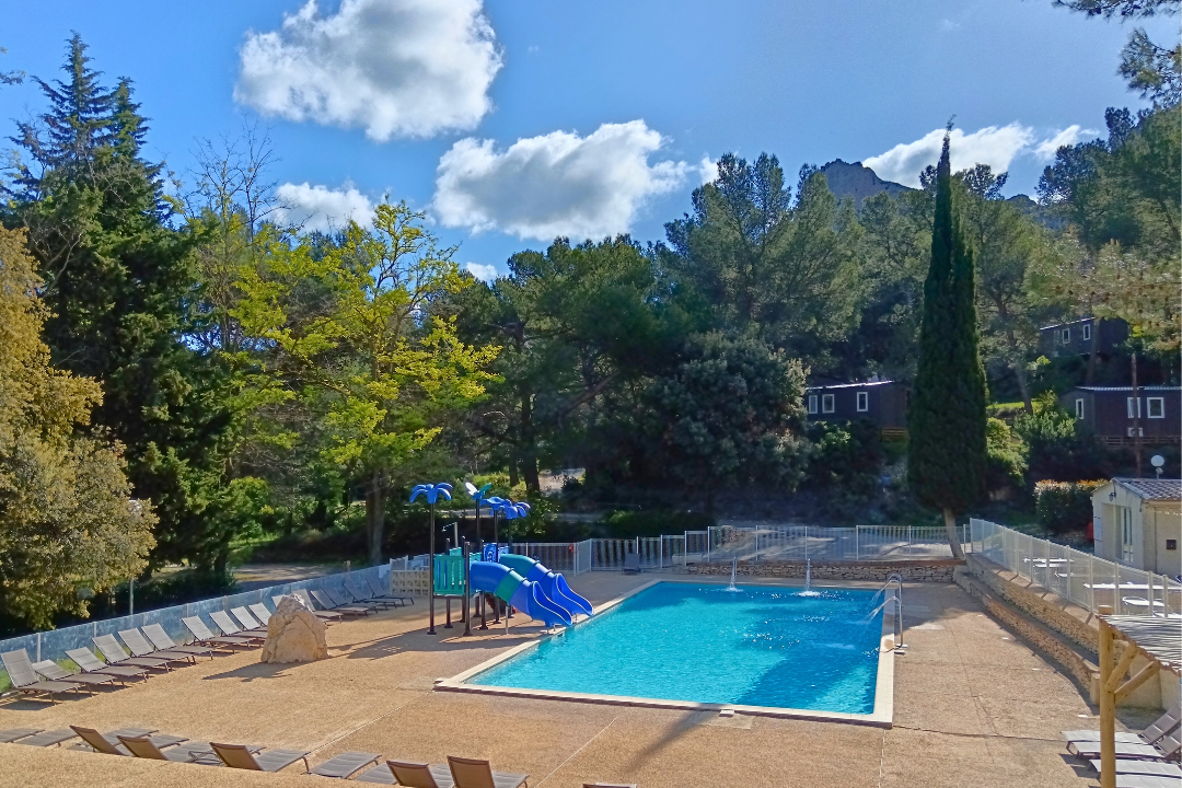 Blaue Camping-Poolanlage mit Umzäunung, Liegeflächen und Spielbereich, im Hintergrund Bäume, Berge und mehrere Gebäude.