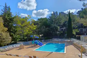 Blaue Camping-Poolanlage mit Umzäunung, Liegeflächen und Spielbereich, im Hintergrund Bäume, Berge und mehrere Gebäude.