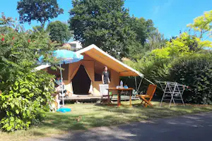 Campingzelt mit Vorzelt und Holzterrasse, Campingstühle und Tisch auf dem Stellplatz im Grünen, umgeben von Bäumen.