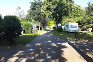 Campingplatz-Allee mit Wohnwagen, Rasenflächen und Hecken, umgeben von Bäumen und einem geparkten Auto am Wegesrand.