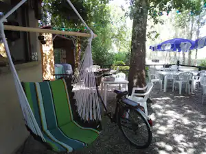 Grüne Camping-Terrasse mit Liegestuhl, Fahrrad und mehreren weißen Stühlen unter Bäumen neben einem Gebäude.