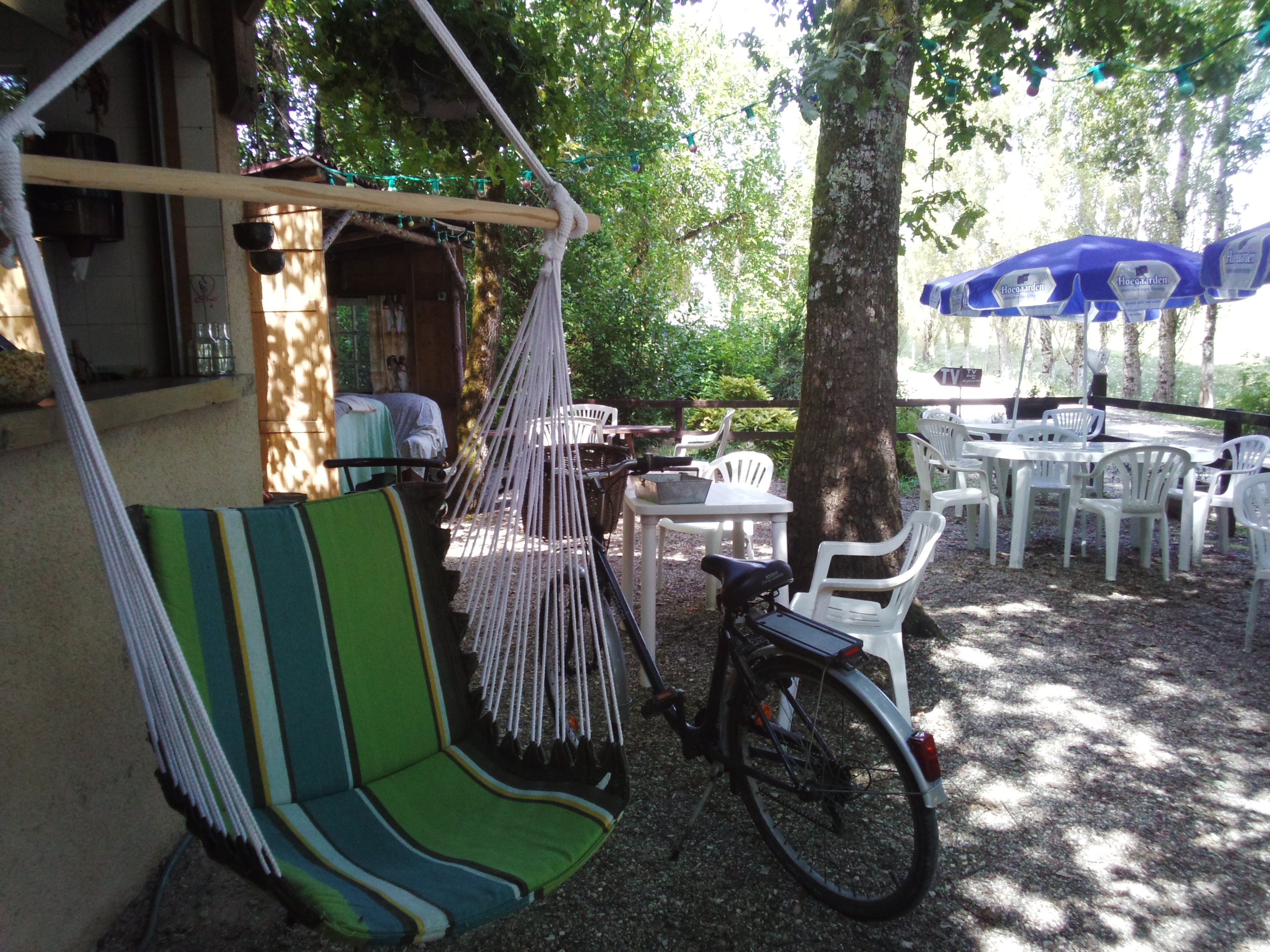 Grüne Sonnenliege auf der Camping-Terrasse, daneben Fahrrad und weiße Stühle unter Bäumen.