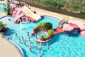 Großes Camping-Schwimmbecken mit Wasserrutsche, Felsattrappen und mehreren Beckenbereichen, umgeben von Grün und Wegen.