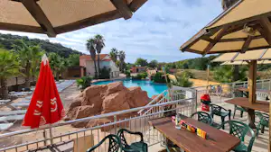 Terrasse mit Sitzbereich und Tisch am Campingplatz, Blick auf den Pool mit Felsattrappe und Palmen im Hintergrund.