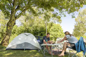 Campingzelt im Grünen unter Bäumen, daneben ein Tisch mit Stühlen, zwei Personen sitzen und essen.
