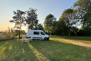 Weißes Wohnmobil auf einem grünen Campingplatz mit Sonnenschirm und Liegestühlen neben dem Fahrzeug, Bäume im Hintergrund.