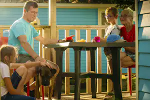 Familie sitzt auf einer Camping-Terrasse, ein Hund liegt daneben, im Hintergrund Holzbalken und bunte Deko-Becher.