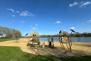 Spielplatz am Seeufer mit Klettergerüst und Schaukeln, Sandfläche und Gras, im Hintergrund Wasser und Bäume.