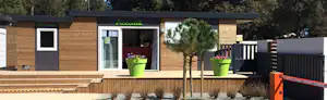Modernes Camping-Haus mit Holzfassade, großen Fenstern und Eingangstür, davor Terrasse mit Sitzmöbeln und Pflanzkübeln.