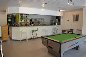 Billardtisch im Vordergrund, dahinter eine Camping-Bar mit Tresen, Getränkeflaschen und Sitzhockern im Innenbereich.