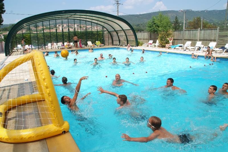 Kinder und Erwachsene im beheizten Pool, im Vordergrund ein gelbes aufblasbares Spielgerät, dahinter Liegen und Zaun.