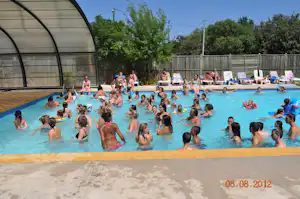 Kinder und Erwachsene im großen Schwimmbecken eines Campingplatzes, daneben Liegen und ein Glasdach, im Hintergrund Bäume und Zaun.