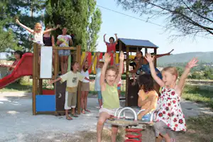 Kinder spielen auf einem Spielplatz mit Rutsche und Klettergerüst, im Hintergrund Bäume und ein Holzhaus.