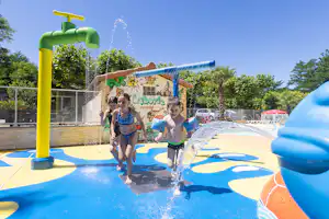 Kinder planschen im Camping-Pool, während Wasser aus einer gelben Fontäne sprudelt und ein blauer Spielbereich im Hintergrund liegt.