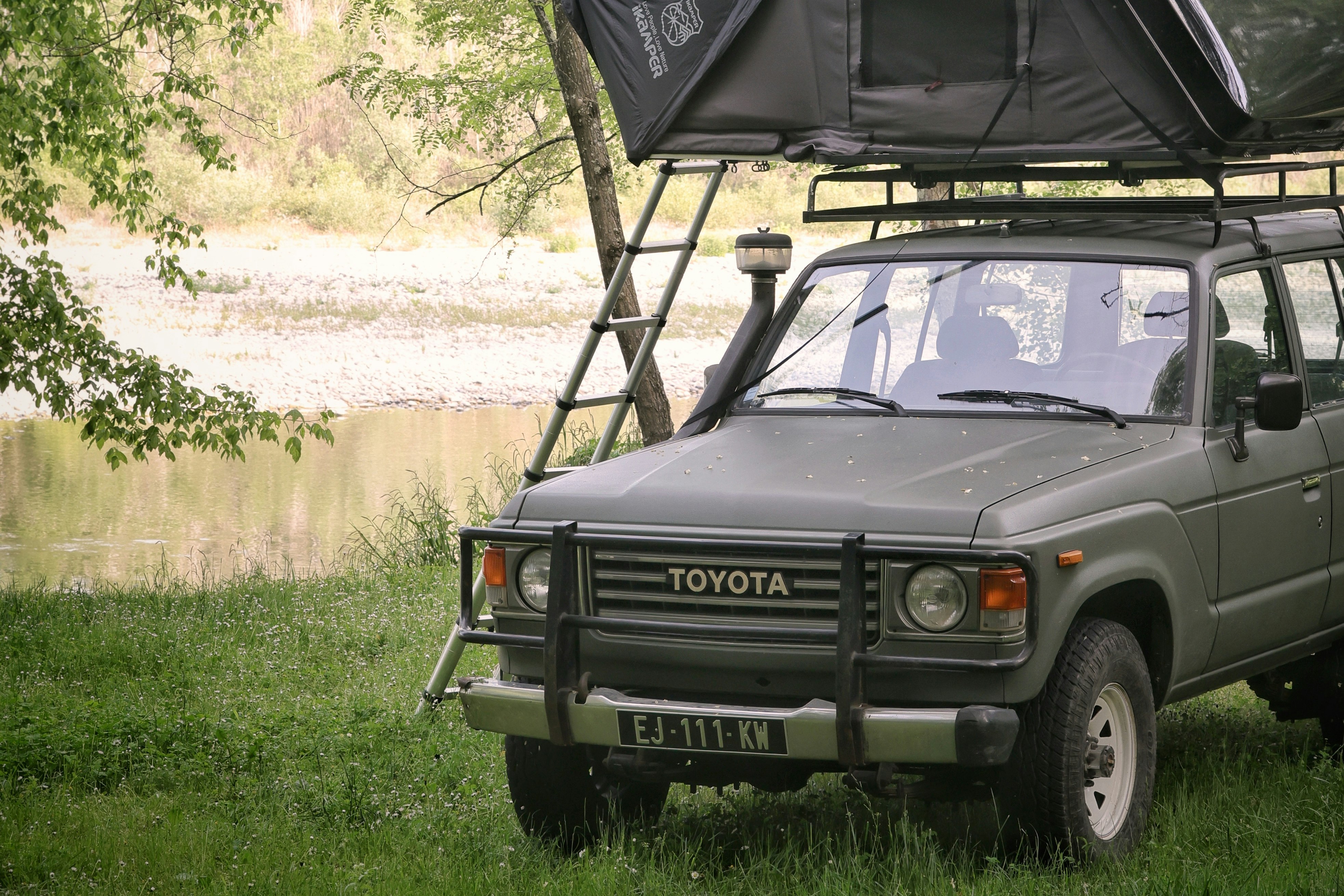 Geländewagen mit Dachzelt auf einem Campingplatz im Grünen, umgeben von Bäumen und Gras, mit Frontscheinwerfern und Kühlergrill.