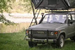 Geländewagen mit Dachzelt auf einem Campingplatz im Grünen, umgeben von Bäumen und Gras, mit Frontscheinwerfern und Kühlergrill.