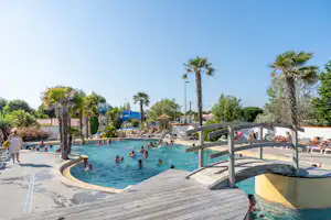 Schwimmbad auf dem Campingplatz mit Palmen, Holzsteg und Liegen, im Hintergrund Gebäude und weitere Gäste im Wasser.
