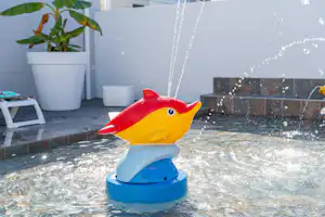 Spielzeug-Fisch auf einem Planschbecken im Pool, Wasser spritzt, weiße Wand und Pflanzkübel im Hintergrund.