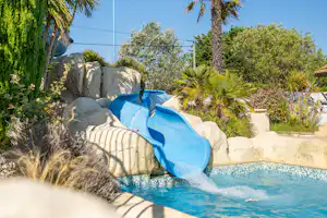 Blauer Pool mit Wasserrutsche, umgeben von Palmen und Sträuchern, Steinmauer und klarem Himmel.
