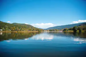 Ruhiger See mit Spiegelung, umgeben von Wäldern und Bergen, im Hintergrund ein bewölkter Himmel.
