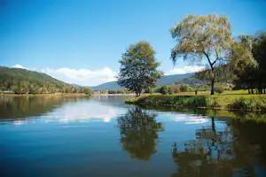 Ruhiger See mit Spiegelung, Bäumen am Ufer und Blick auf bewaldete Hügel und Berge in der Ferne.